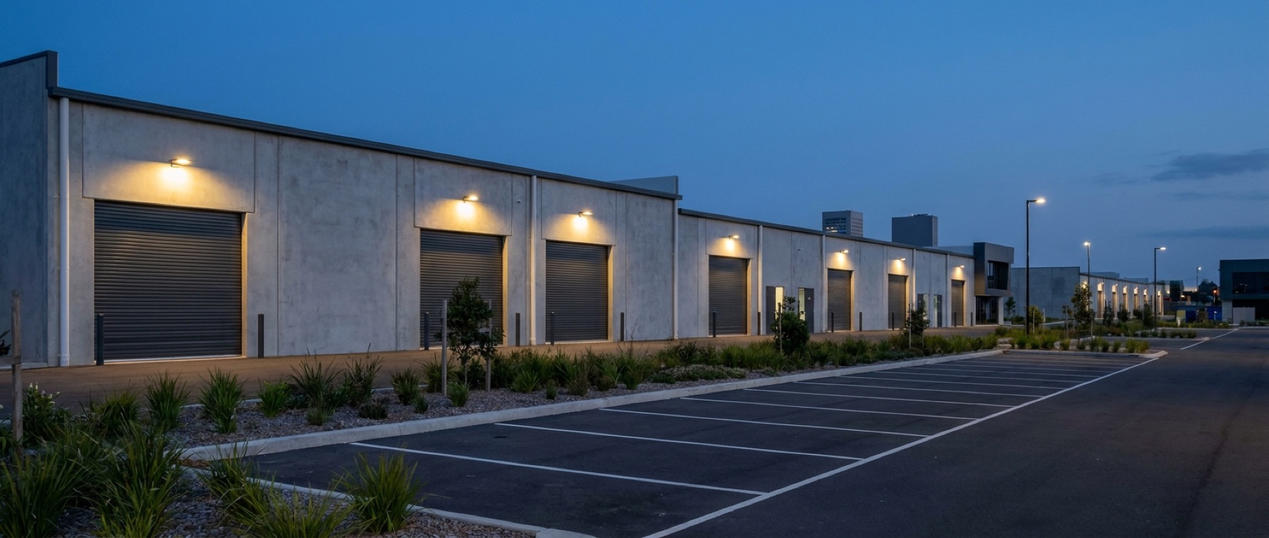 A row of small workshop units in an industrial estate at dusk, roller shutters half lowered, one service light burning above an open unit.